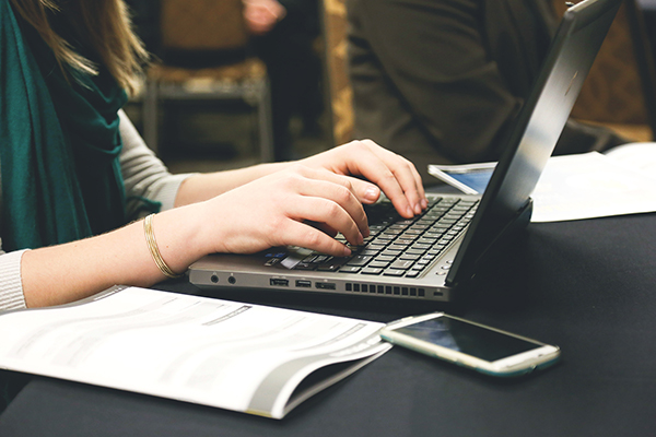 woman typing on a laptop computer on a desk with an open notebook and mobile phone beside her